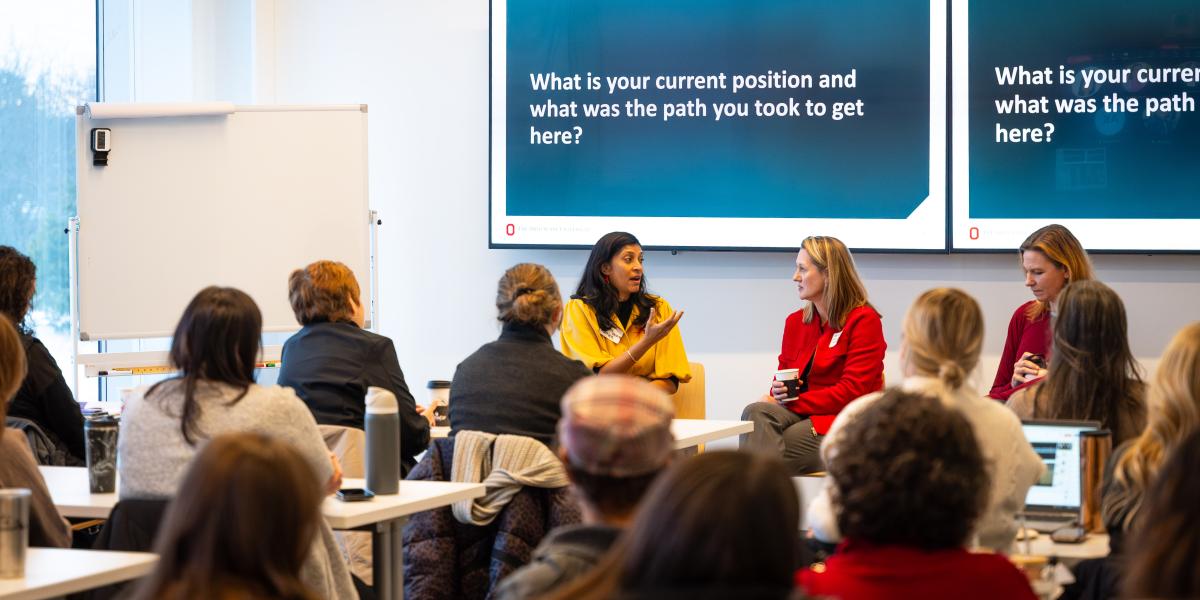 A group of women are seen sitting listening to three women speak who are seated at the front of a room.