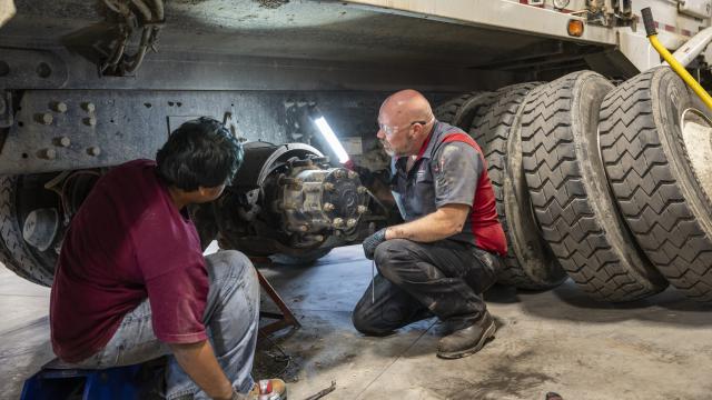 Two mechanics use a light to work on the bottom of a bus.